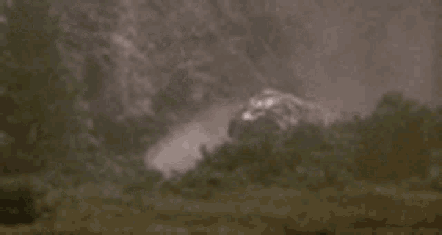a helicopter is flying over a field with trees in the background and smoke coming out of it .