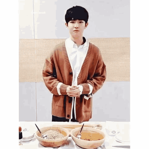 a young man in a brown cardigan is standing in front of a table with bowls of food on it .