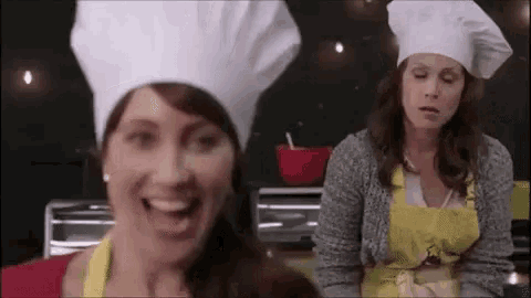 two women wearing chef hats and aprons are smiling in a kitchen