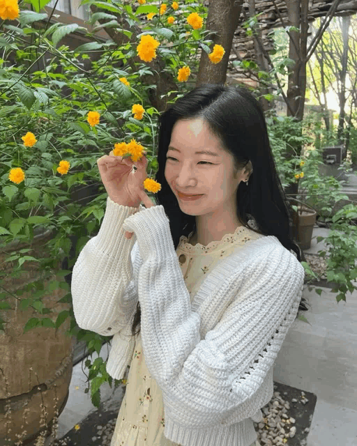 a woman wearing a white sweater and a white dress smells a yellow flower