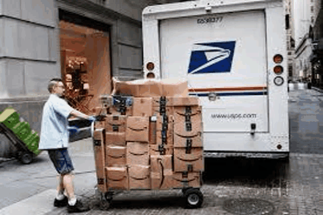 a man is pushing a cart filled with boxes in front of a usps truck .