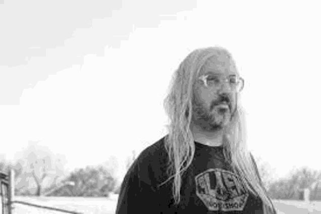 a black and white photo of a man with long blonde hair and a beard wearing glasses .