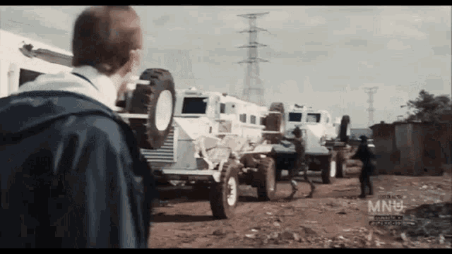 a man in a blue jacket stands in front of a row of military vehicles with the words mnu on the bottom