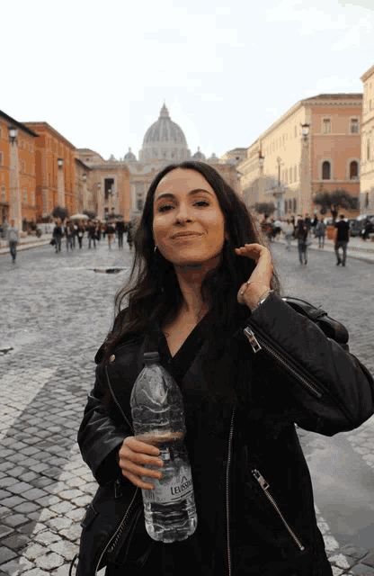 a woman in a black leather jacket holds a bottle of l' acqua in her hand