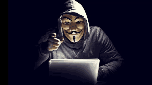 a man wearing an anonymous mask is pointing at a laptop screen