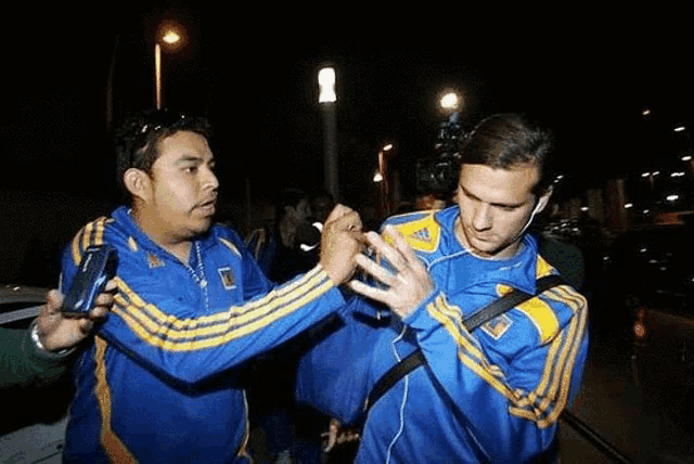 a man wearing a blue and yellow adidas shirt talks to another man