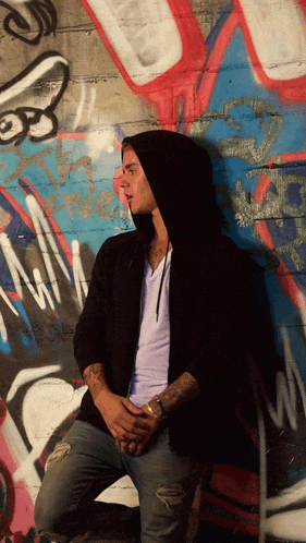 a man in a black hoodie is kneeling in front of a graffiti wall