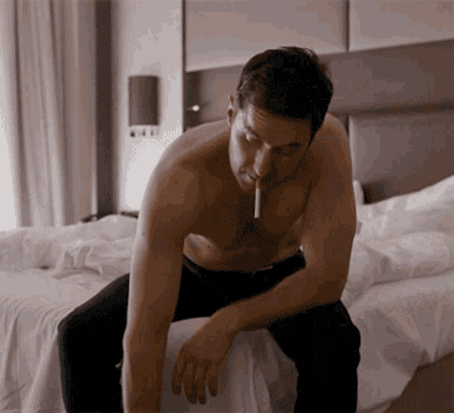 a shirtless man with a cigarette in his mouth sits on a bed