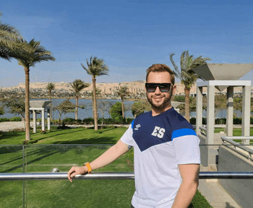 a man wearing sunglasses and a shirt that says es stands in front of a lake