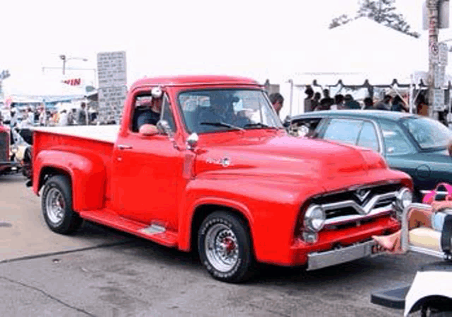 a red pickup truck is parked in front of a crowd of people .