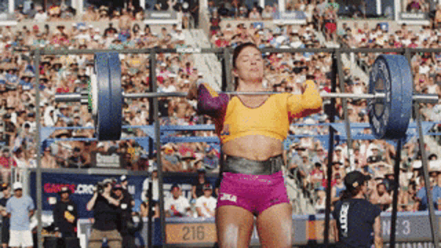 a woman is lifting a barbell in front of a crowd and the number 216 is on the wall behind her