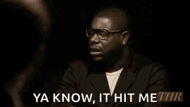a man wearing glasses is sitting in a dark room and talking to another person .