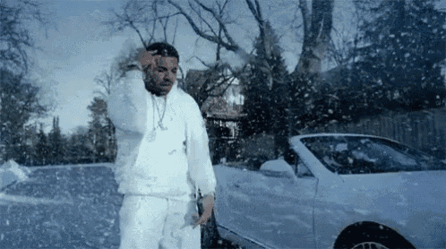 a man in a white jacket and white pants is standing in front of a white car in the snow .