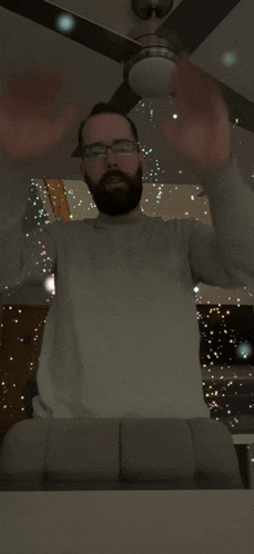 a man with a beard and glasses stands in front of a ceiling fan