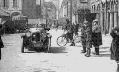 a black and white photo of a car driving down a city street with people standing on the sidewalk .