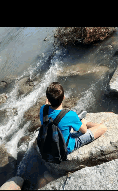 a boy with a backpack sits on a rock near a river