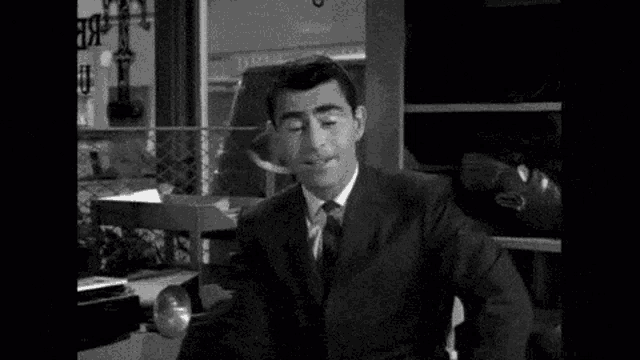 a man in a suit and tie is smoking a cigarette while sitting at a table in a black and white photo .