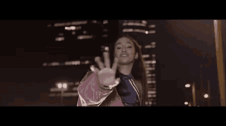 a woman is standing in front of a city skyline at night and making a stop sign with her hand .