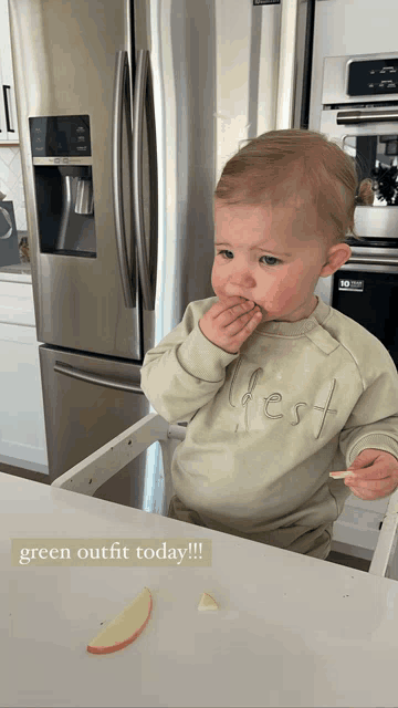 a baby wearing a green outfit is eating an apple slice