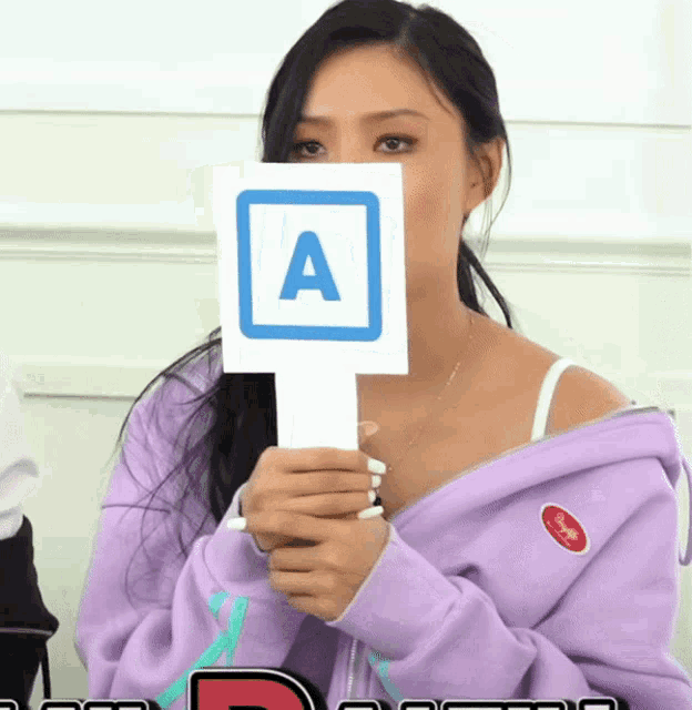 a woman in a purple jacket holds a sign with the letter a on it in front of her face