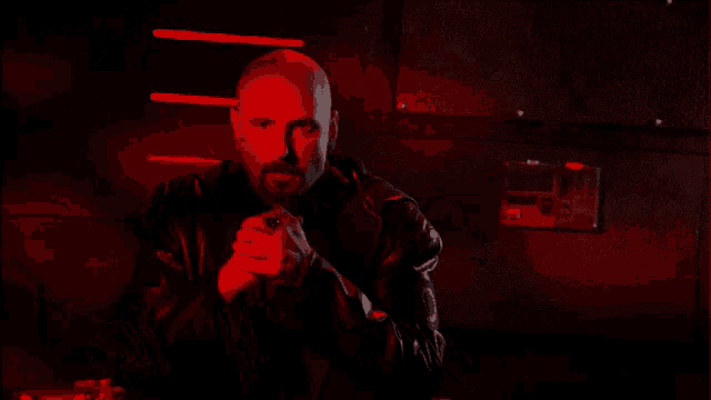 a bald man with a beard wearing a black leather jacket in a dark room with red lights behind him