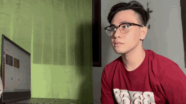 a man wearing glasses and a red shirt with the number 36 on it looks at a laptop