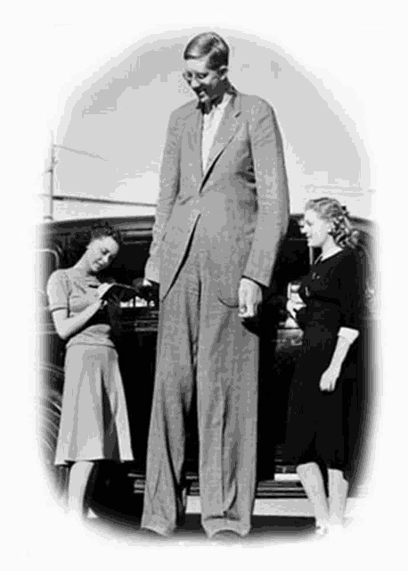 a black and white photo of a very tall man standing next to two women