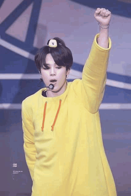 jimin of bts is wearing a yellow sweater and a yellow hat while holding his fist up in the air .