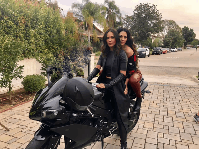 two women are sitting on a black motorcycle in front of a street