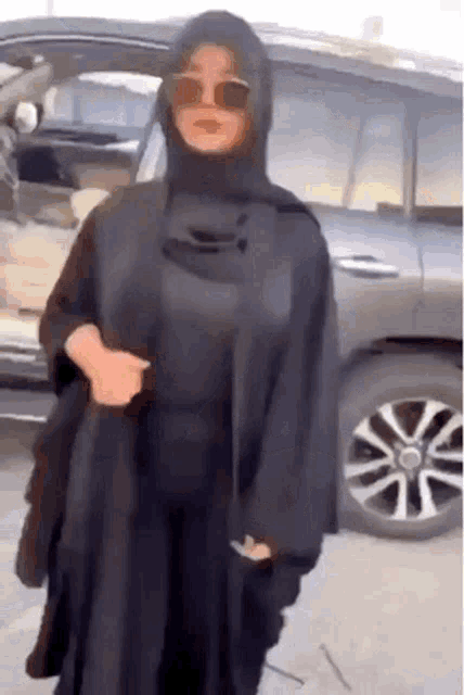 a woman wearing a hijab and sunglasses stands in front of a car .