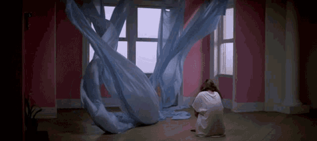 a woman in a white dress sits on the floor in front of a window with blue curtains hanging from it
