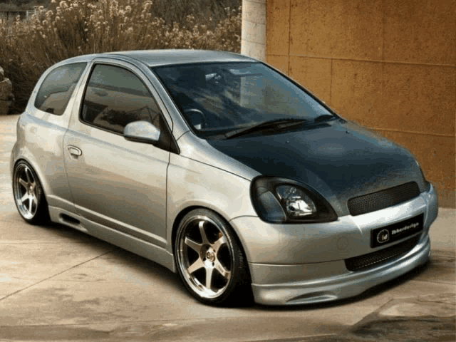 a silver car with a black hood is parked in front of a brown wall