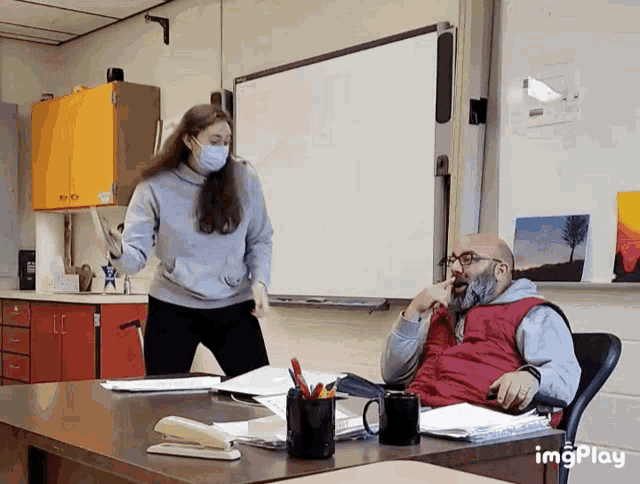 a man sits at a desk in front of a white board while a woman stands behind him wearing a mask