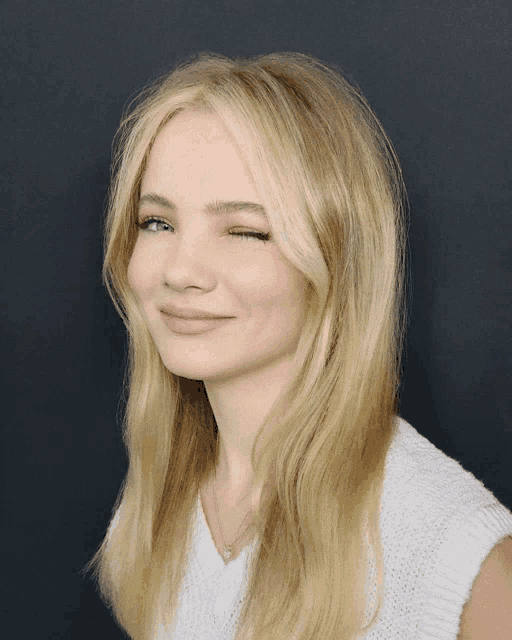 a woman with blonde hair and a white sweater winks at the camera