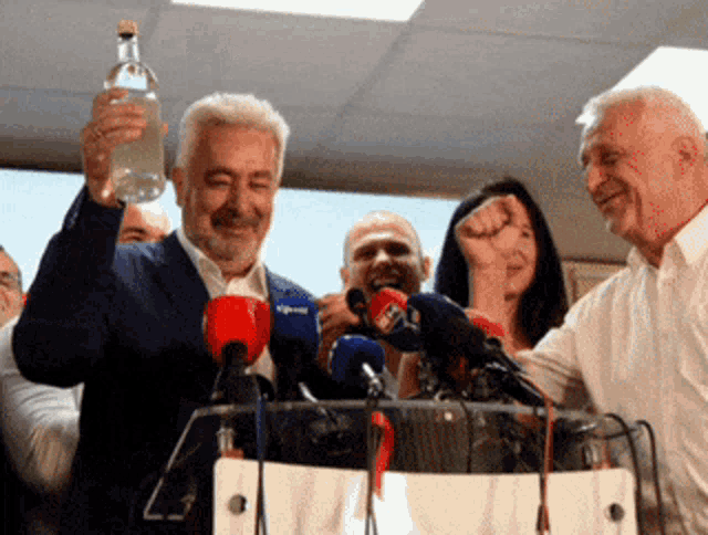 a man holds up a bottle of water in front of a group of microphones including one that says rtc