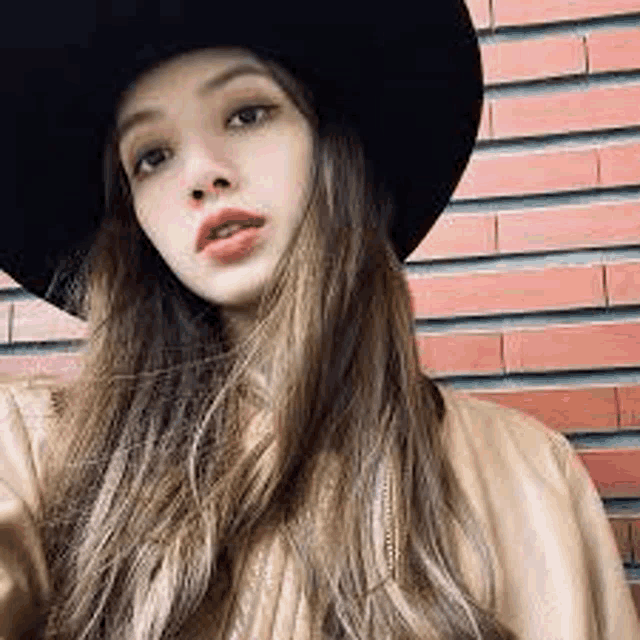 a woman wearing a hat and a leather jacket is standing in front of a red brick wall .