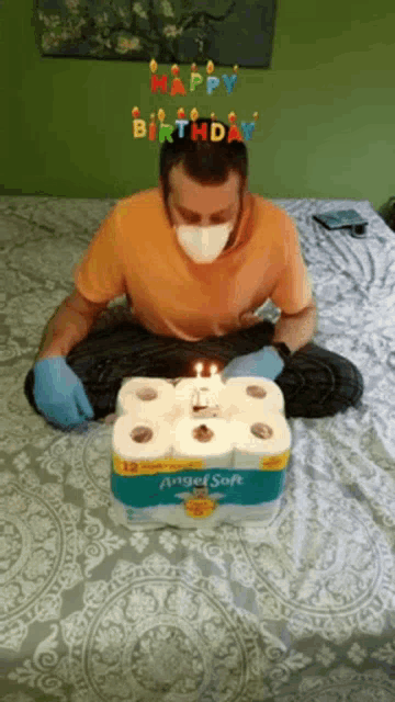 a man wearing a mask and gloves is sitting on a bed next to a birthday cake made out of toilet paper rolls .