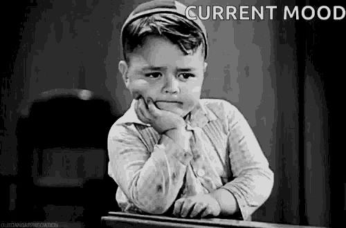 a little boy is sitting at a desk with his hand on his chin and making a sad face .