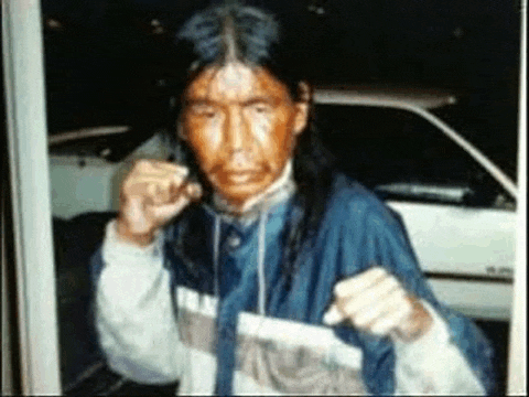 a man with long hair and a blue jacket is standing in front of a car .