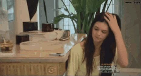 a woman is sitting at a table with her hand on her head and a plant in the background .