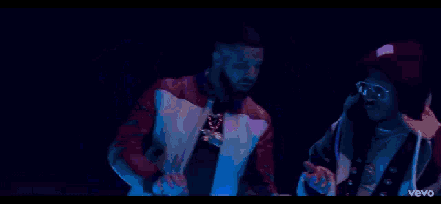 two men are standing next to each other in a dark room . one of the men is wearing a hat .