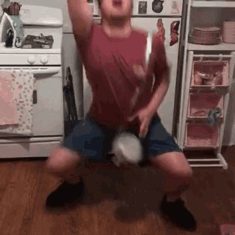 a man in a red shirt and blue shorts is squatting down in front of a stove and refrigerator .