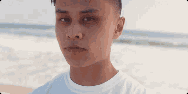 a young man in a white shirt stands on the beach looking at the camera