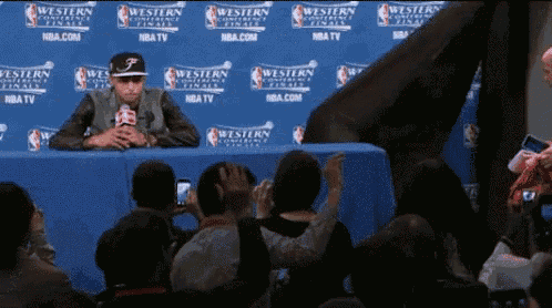 a man sitting at a table with a microphone in front of a wall that says western conference finals on it