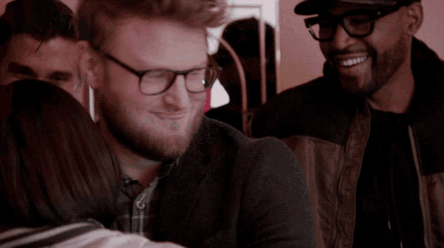 a man wearing glasses and a hat smiles while a woman hugs him