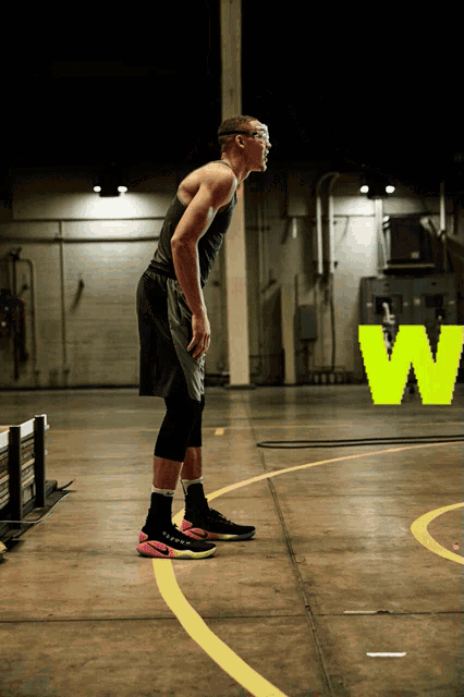 a man in a black tank top and black shorts stands on a basketball court in front of a neon w sign