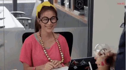 a woman wearing glasses and a pearl necklace is sitting at a desk in front of a laptop computer .