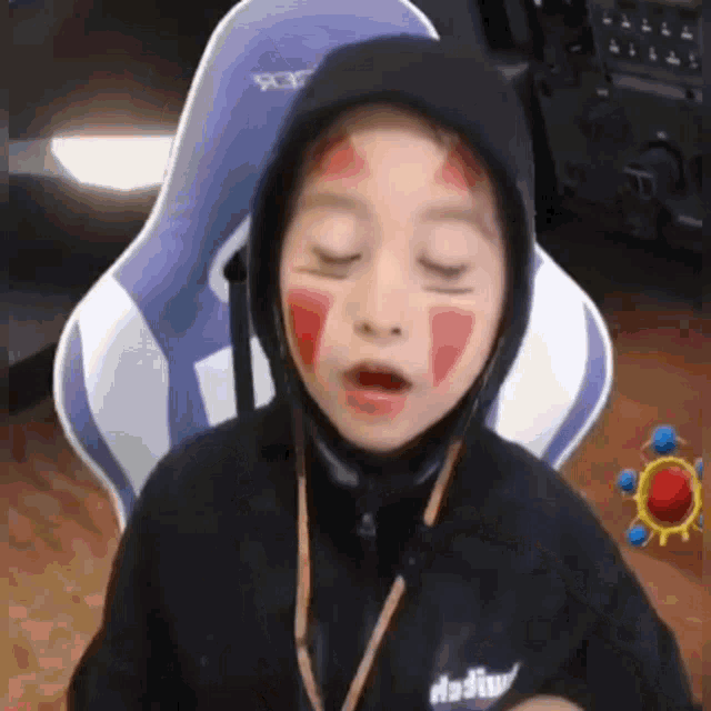 a young boy with red paint on his face is wearing a hooded jacket and sitting in a chair .