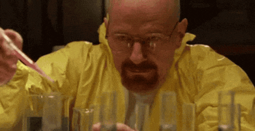 a man in a yellow suit is sitting at a table with beakers and a pipette in his hand .