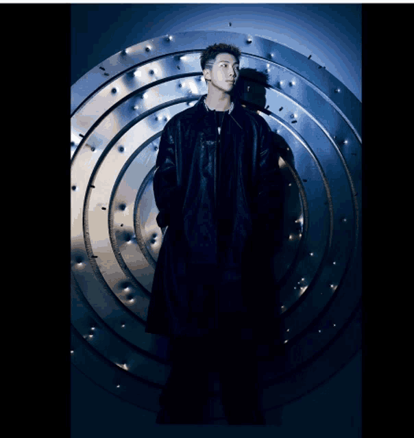 a man in a black coat stands in front of a metal wall with holes in it